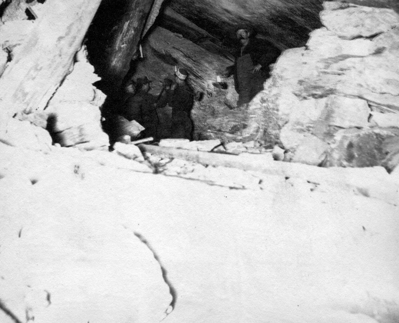 A group of men wearing long pants and shirts, one carrying a lamp, enters one of the small and dark mine shafts at Ely, being supported by wooden poles and piles of rocks. | UVM Landscape Change Program 